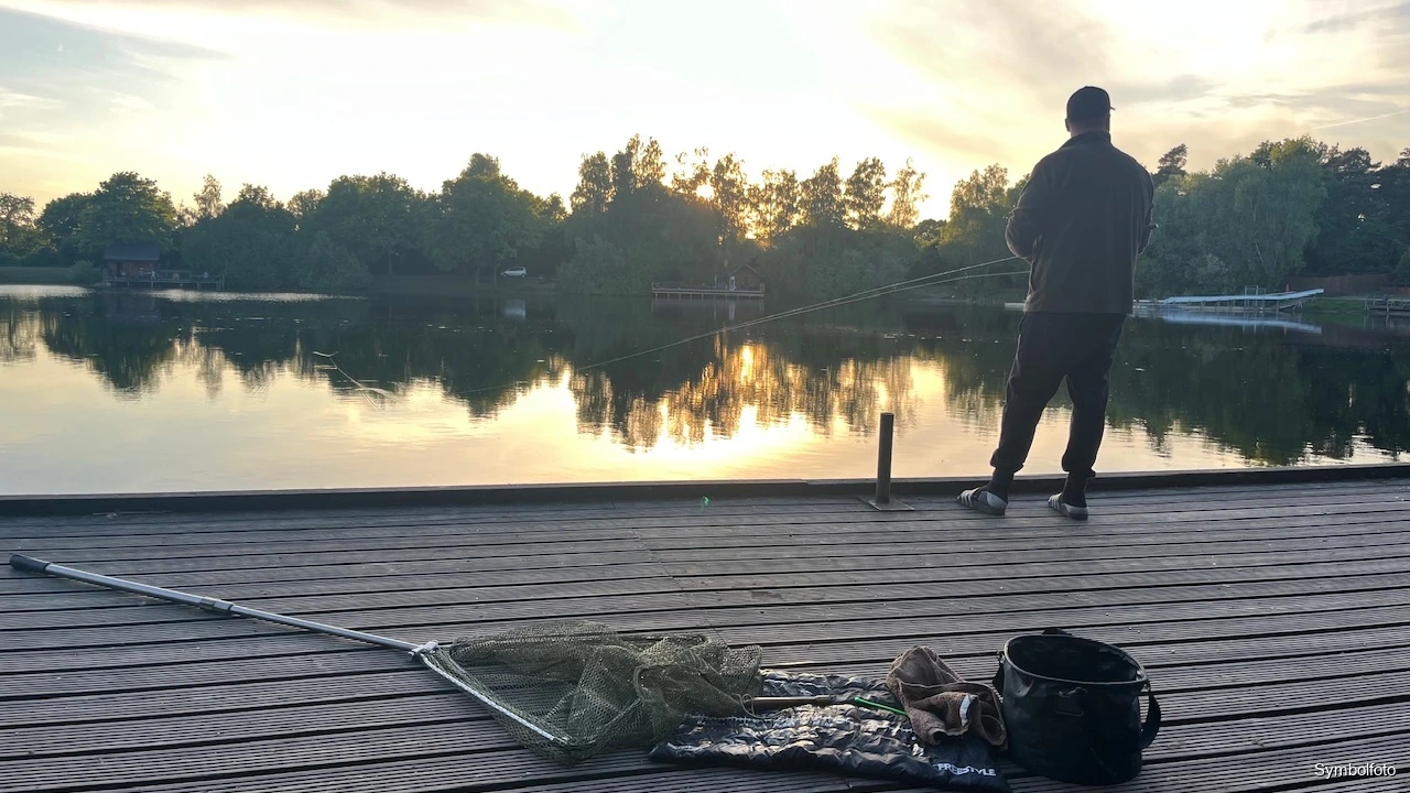 Angler am Holzsteg beim Ansitz in der Abenddämmerung an einem Forellensee, Kescher im Vordergrund